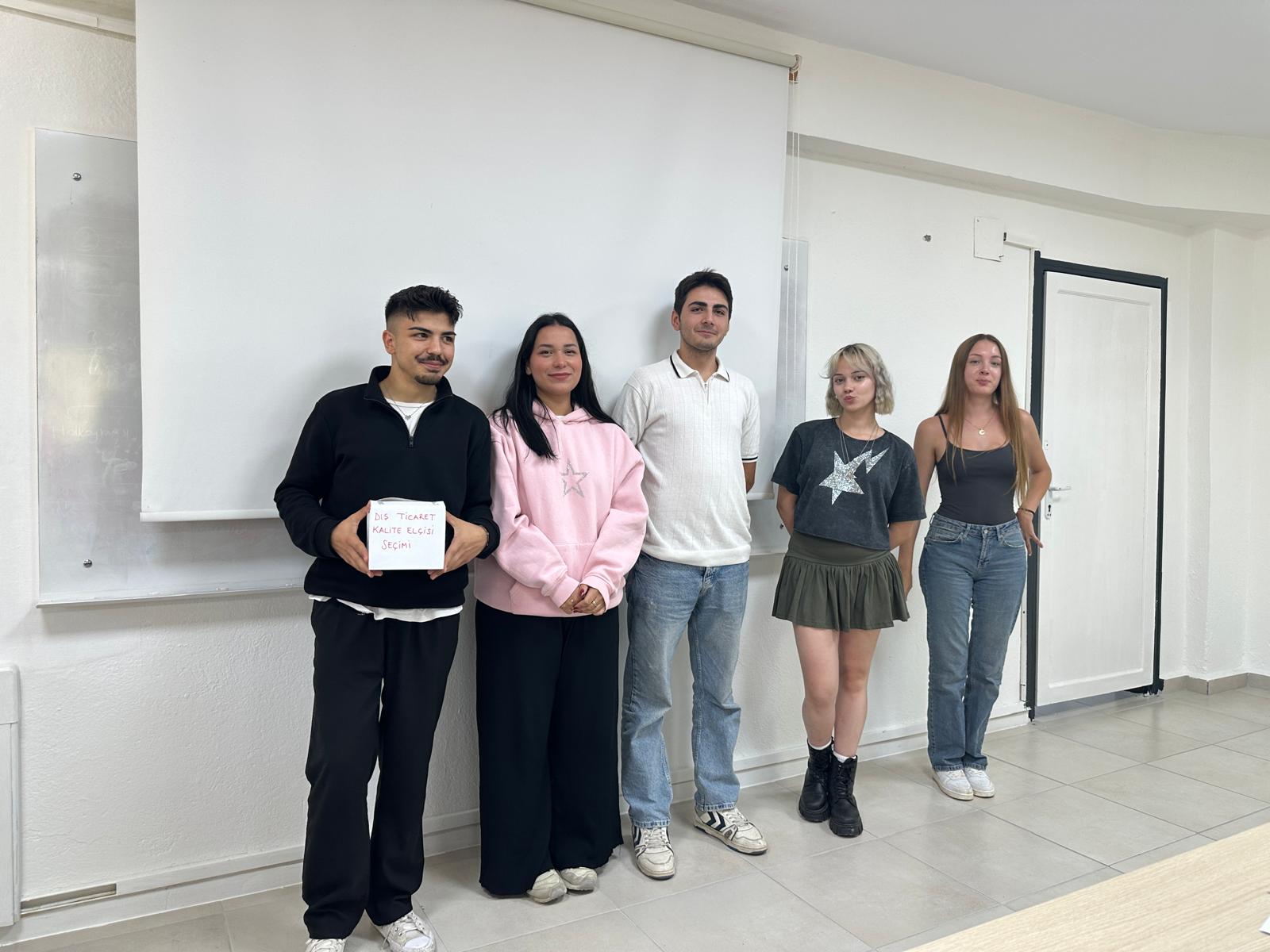 In a classroom setting, five university students are standing side by side. Behind them is a white projection screen and a whiteboard.  * The student on the far left is wearing a black tracksuit and holding a white box labeled “Foreign Trade Quality Ambassador Election” in red letters.  * Next to him, a student in a pink hoodie is smiling warmly.  * The middle student is wearing a white polo shirt and blue jeans.  * The student next to him has a gray t-shirt with a star print, a green mini skirt, and black boots.  * The student on the far right is dressed in a gray tank top and blue jeans, with long hair. All five stand in front of a plain white wall, posing in a friendly group photo.